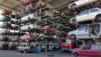 Numerous stacked rows of disassembled cars are placed on metal structures in what appears to be a scrapyard. Many of the cars are missing parts like wheels and doors.