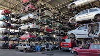 Numerous stacked rows of disassembled cars are placed on metal structures in what appears to be a scrapyard. Many of the cars are missing parts like wheels and doors.