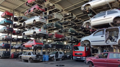 Numerous stacked rows of disassembled cars are placed on metal structures in what appears to be a scrapyard. Many of the cars are missing parts like wheels and doors.