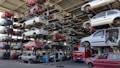 Numerous stacked rows of disassembled cars are placed on metal structures in what appears to be a scrapyard. Many of the cars are missing parts like wheels and doors.