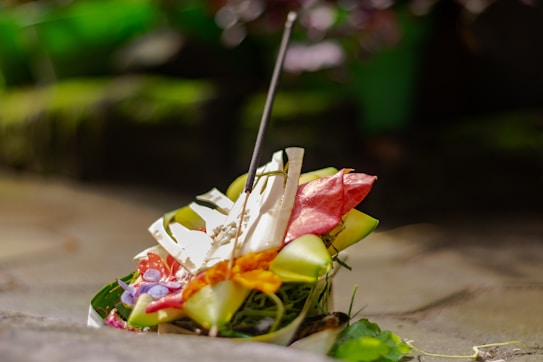 An offering made from various natural elements, including colorful flowers, leaves, and a stick resembling incense, placed on a stone surface. The setting appears calm and serene, with blurred green foliage in the background adding to the tranquil atmosphere.