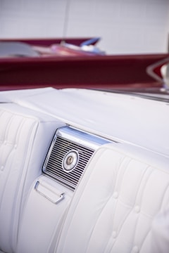 A detailed view of the interior of a car highlighting a section of a white leather seat with an elegant design. The seat features intricate stitching and a built-in air vent with a metallic finish. The background shows a hint of burgundy, possibly another part of the car interior or a convertible roof.