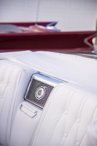 A detailed view of the interior of a car highlighting a section of a white leather seat with an elegant design. The seat features intricate stitching and a built-in air vent with a metallic finish. The background shows a hint of burgundy, possibly another part of the car interior or a convertible roof.