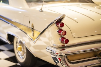 A vintage car featuring a cream-colored exterior with shiny chrome detailing. The rear fin showcases three classic red tail lights stacked vertically. The reflections and lighting highlight its polished surface, with a gold accent running along the side.