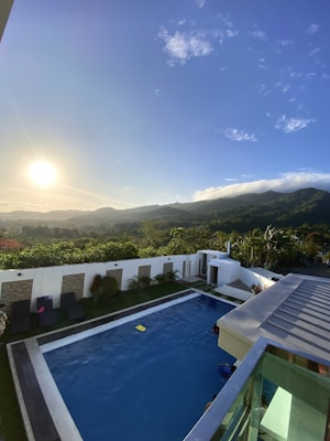 A picturesque landscape featuring a swimming pool surrounded by lush greenery and mountains in the distance. The sun is setting, casting a golden glow over the scene, with a partially cloudy sky above. Outdoor lounge chairs line one side of the pool, and a small structure is located at the far end, near the foliage.