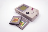 A vintage handheld gaming console with a gray body, featuring a rectangular design, a monochrome screen, and black and magenta buttons is displayed alongside two game cartridges. The cartridges have distinct labels, suggesting different titles.