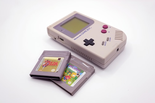 A vintage handheld gaming console with a gray body, featuring a rectangular design, a monochrome screen, and black and magenta buttons is displayed alongside two game cartridges. The cartridges have distinct labels, suggesting different titles.