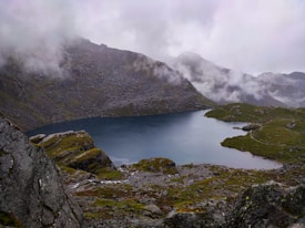 A serene mountain lake nestled among rugged peaks and rocky terrain. Misty clouds hover above, partially shrouding the mountains in the background. The foreground features moss-covered rocks and patches of greenery, creating a tranquil and untouched atmosphere.