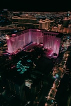 Aerial view of a cityscape at night, prominently displaying a large hotel with pink and purple lighting. The surrounding area is illuminated with colorful lights and includes various buildings and structures. Streets are lined with lit pathways and signs, enhancing the vibrant urban scene.