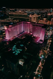 Aerial view of a cityscape at night, prominently displaying a large hotel with pink and purple lighting. The surrounding area is illuminated with colorful lights and includes various buildings and structures. Streets are lined with lit pathways and signs, enhancing the vibrant urban scene.
