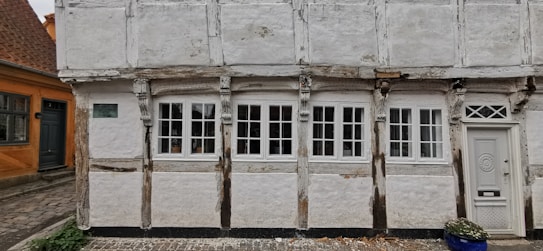 A traditional half-timbered house with white plaster and exposed wooden beams, featuring five multi-paned windows and a decorative wooden door with intricate designs. The house appears weathered, adding to its historic charm. There is a cobbled path in front, and part of a blue ceramic pot with a plant can be seen to the right.