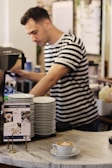 A barista wearing a striped shirt prepares a coffee drink in a café. There is a stack of white plates on the counter and a small sign with information about workshop tariffs. In the foreground, a cappuccino with latte art sits on a marble counter.