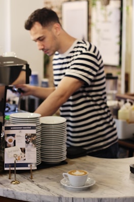 A barista wearing a striped shirt prepares a coffee drink in a café. There is a stack of white plates on the counter and a small sign with information about workshop tariffs. In the foreground, a cappuccino with latte art sits on a marble counter.