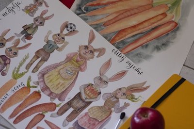 The image features an illustration of anthropomorphic rabbits dressed in various outfits, such as a soccer jersey and an apron. Some are holding objects like a basket and a soccer ball. The artwork includes depictions of orange carrots and handwritten text in a foreign language. Accompanying the illustration are art supplies, including a paintbrush and a yellow notebook, with a red apple placed on the side.