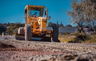 A large, yellow construction vehicle with thick tires is working on a road under a clear blue sky. The machine appears to be a grader or a road roller, actively leveling or compacting the dirt. The surrounding area includes trees and buildings in the background, indicating an urban or suburban setting.