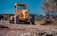 A large, yellow construction vehicle with thick tires is working on a road under a clear blue sky. The machine appears to be a grader or a road roller, actively leveling or compacting the dirt. The surrounding area includes trees and buildings in the background, indicating an urban or suburban setting.