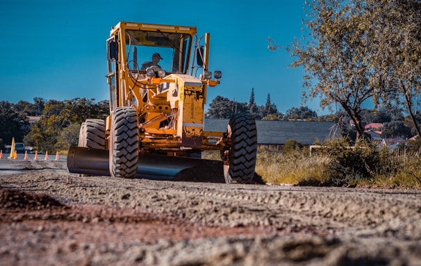 A large, yellow construction vehicle with thick tires is working on a road under a clear blue sky. The machine appears to be a grader or a road roller, actively leveling or compacting the dirt. The surrounding area includes trees and buildings in the background, indicating an urban or suburban setting.