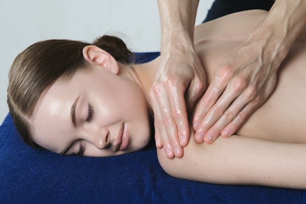 A woman is lying on a blue towel with her eyes closed, receiving a back massage. The hands of the person giving the massage are visible, applying gentle pressure to the woman's upper back.