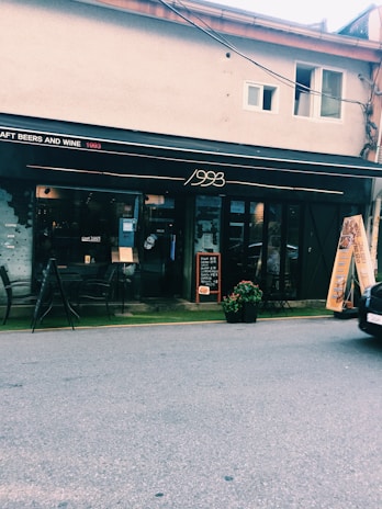 A street view of a cafe named '1993', featuring a black sign with white and red lettering advertising craft beers and wine. The storefront has a modern, minimalist design with glass windows and a few patio chairs and tables outside. A chalkboard menu and a potted plant are near the entrance. The street is lightly trafficked with visible road and part of a car.