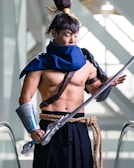A person dressed in a detailed costume, resembling a warrior or samurai character, complete with a blue cape, arm guards, and a large sword. The outfit is accessorized with a rope belt, a topknot hairstyle, and leather straps. The setting appears to be indoors, with escalator railings and natural light coming through large windows in the background.