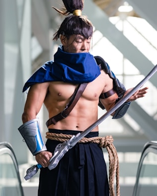 A person dressed in a detailed costume, resembling a warrior or samurai character, complete with a blue cape, arm guards, and a large sword. The outfit is accessorized with a rope belt, a topknot hairstyle, and leather straps. The setting appears to be indoors, with escalator railings and natural light coming through large windows in the background.