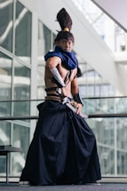 A person dressed in elaborate cosplay attire stands in a poised stance inside a modern building with large glass windows. The costume includes a long, dark skirt-like garment, a blue scarf, and arm guards. The individual also has a distinctive high ponytail hairstyle.