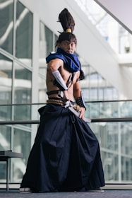 A person dressed in elaborate cosplay attire stands in a poised stance inside a modern building with large glass windows. The costume includes a long, dark skirt-like garment, a blue scarf, and arm guards. The individual also has a distinctive high ponytail hairstyle.