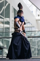 A person dressed in elaborate cosplay attire stands in a poised stance inside a modern building with large glass windows. The costume includes a long, dark skirt-like garment, a blue scarf, and arm guards. The individual also has a distinctive high ponytail hairstyle.