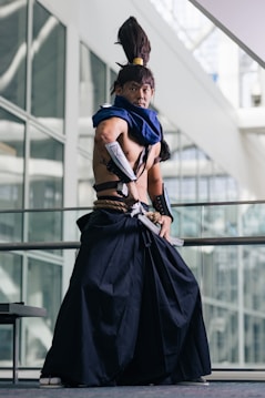 A person dressed in elaborate cosplay attire stands in a poised stance inside a modern building with large glass windows. The costume includes a long, dark skirt-like garment, a blue scarf, and arm guards. The individual also has a distinctive high ponytail hairstyle.