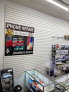 A retail store interior featuring a poster for phone repair, showcasing various smartphone images and the slogan 'You Break It, We Fix It!'. An ATM machine is positioned nearby, and a glass display case contains various products such as watches and small electronic items. Shelves under the counter hold boxed products.