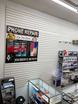A retail store interior featuring a poster for phone repair, showcasing various smartphone images and the slogan 'You Break It, We Fix It!'. An ATM machine is positioned nearby, and a glass display case contains various products such as watches and small electronic items. Shelves under the counter hold boxed products.