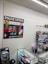 A retail store interior featuring a poster for phone repair, showcasing various smartphone images and the slogan 'You Break It, We Fix It!'. An ATM machine is positioned nearby, and a glass display case contains various products such as watches and small electronic items. Shelves under the counter hold boxed products.