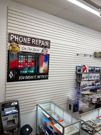 A retail store interior featuring a poster for phone repair, showcasing various smartphone images and the slogan 'You Break It, We Fix It!'. An ATM machine is positioned nearby, and a glass display case contains various products such as watches and small electronic items. Shelves under the counter hold boxed products.
