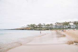 A sandy beach with gentle waves, where people are enjoying leisurely activities. The shoreline curves to the right, and a backdrop of lush greenery and palm trees separates the beach from large resort buildings. The sky is overcast, contributing to a soft and calm atmosphere.