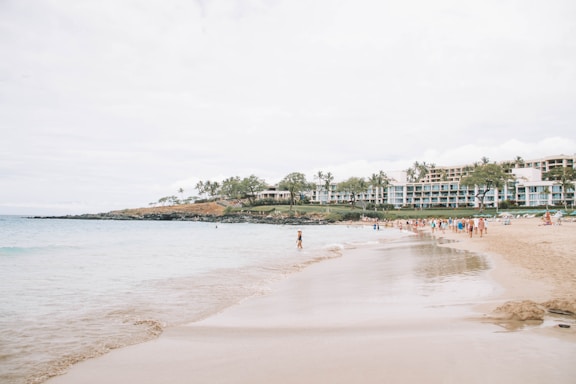 A sandy beach with gentle waves, where people are enjoying leisurely activities. The shoreline curves to the right, and a backdrop of lush greenery and palm trees separates the beach from large resort buildings. The sky is overcast, contributing to a soft and calm atmosphere.