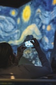 A person holds a smartphone, capturing a photo of a projected art piece that resembles the swirling starry sky theme of Van Gogh’s Starry Night. The background features vivid blue and yellow swirls, creating a dynamic and immersive art experience.