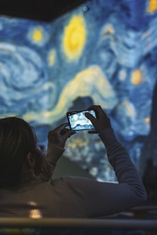 A person holds a smartphone, capturing a photo of a projected art piece that resembles the swirling starry sky theme of Van Gogh’s Starry Night. The background features vivid blue and yellow swirls, creating a dynamic and immersive art experience.