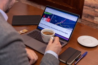 A person in a suit holding a cup of coffee sits at a wooden table with a laptop displaying a cryptocurrency price chart and news headline. Nearby are a notepad with a pen, a tablet, and two coins, suggesting a workspace focused on financial activities.