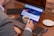 A person in a suit holding a cup of coffee sits at a wooden table with a laptop displaying a cryptocurrency price chart and news headline. Nearby are a notepad with a pen, a tablet, and two coins, suggesting a workspace focused on financial activities.