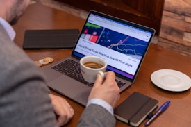 A person in a suit holding a cup of coffee sits at a wooden table with a laptop displaying a cryptocurrency price chart and news headline. Nearby are a notepad with a pen, a tablet, and two coins, suggesting a workspace focused on financial activities.