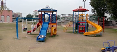 A playground equipped with a variety of play structures including slides, swings, and climbing frames. A blue slide extends from one structure, while a red and yellow spiral slide curves down from another. Swings are positioned to the left, and in the background, there is a small bridge and a few buildings.