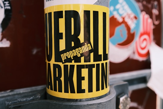 A street pole features a bold yellow and black poster with the words 'Guerilla Marketing' and 'propaganda'. Surrounding the pole are other urban signs with varied colors and designs, hinting at a busy city environment.
