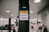 A sign is taped to a pillar in a modern, spacious indoor area. The sign features the colors blue and yellow, resembling the Ukrainian flag, and contains Ukrainian text. The background shows an escalator, some fencing, and a few individuals standing, suggesting a public or transitional space like a station or an airport.