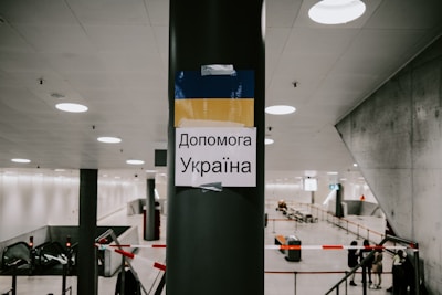 A sign is taped to a pillar in a modern, spacious indoor area. The sign features the colors blue and yellow, resembling the Ukrainian flag, and contains Ukrainian text. The background shows an escalator, some fencing, and a few individuals standing, suggesting a public or transitional space like a station or an airport.