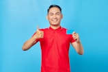 A person wearing a bright red polo shirt, standing against a solid blue background, gives a thumbs-up with one hand and holds a blue card with the other hand. The person is smiling and appears to be happy or content.