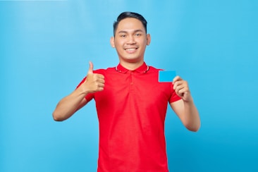 A person wearing a bright red polo shirt, standing against a solid blue background, gives a thumbs-up with one hand and holds a blue card with the other hand. The person is smiling and appears to be happy or content.