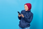 A woman wearing a red hijab and a navy blue fuzzy jacket is looking at her phone with a joyful expression against a bright blue background. Her hand is poised mid-interaction with the phone, indicating engagement with the device.