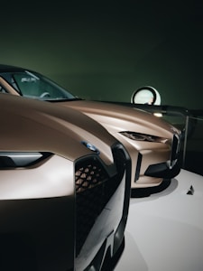 Two sleek, modern cars with smooth, aerodynamic curves and a metallic bronze finish are showcased. The focus is on the front grilles and headlights, highlighting the intricate design and sharp lines. The setting appears to be a showroom with dim lighting, enhancing the luxury feel of the vehicles.