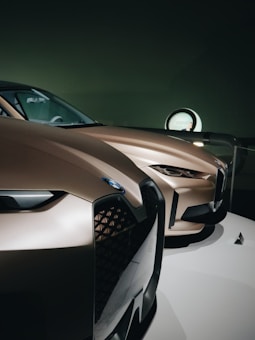 Two sleek, modern cars with smooth, aerodynamic curves and a metallic bronze finish are showcased. The focus is on the front grilles and headlights, highlighting the intricate design and sharp lines. The setting appears to be a showroom with dim lighting, enhancing the luxury feel of the vehicles.