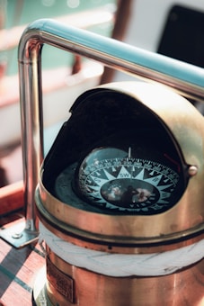 A brass compass housed in a polished casing is mounted on a wooden surface. The compass face displays cardinal and ordinal directions, with an arrow pointing towards the southwest. The surrounding area includes metal railings, and soft natural light highlights the craftsmanship of the object.